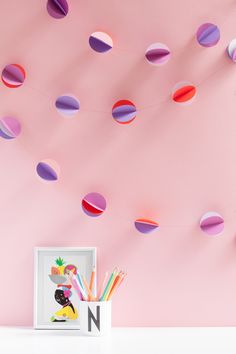 a pink wall with colorful paper circles hanging from it's ceiling and a white frame holding pencils