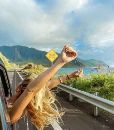 a woman holding her arm out the window of a car on a road with mountains in the background