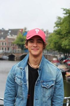 a young man wearing a pink hat standing in front of a river with boats on it