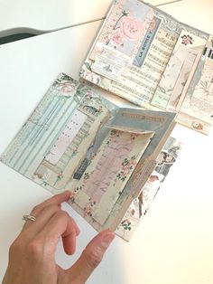 a person holding an open book on top of a white table next to some papers