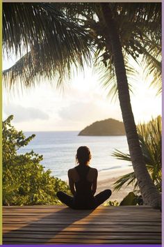 a woman is sitting on the ground in front of some palm trees and looking out at the ocean