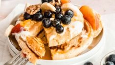 pancakes with blueberries, bananas and other toppings on a white plate next to silverware