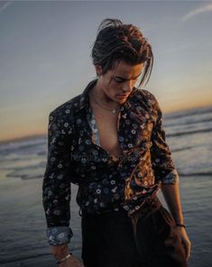 a woman standing on the beach at sunset with her hands in her pockets and looking down