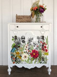 a white cabinet with flowers painted on the front and bottom, next to a basket full of flowers