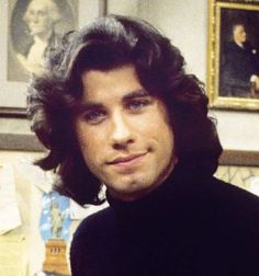 a man with long hair wearing a black turtle neck sweater in front of some paintings