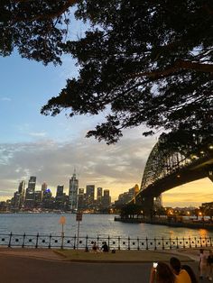 people are walking along the water near a bridge and some buildings in the background at sunset