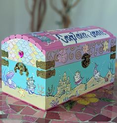 a decorative box sitting on top of a pink and blue tablecloth covered surface with an image of mermaids