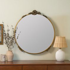 a round mirror sitting on top of a wooden dresser next to a lamp and vase