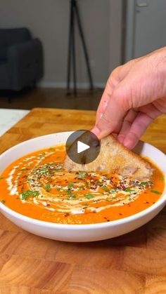 a person dipping a piece of bread into a bowl of tomato soup on a wooden table