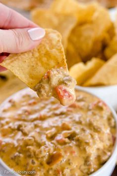 a hand holding a tortilla chip over a bowl of chili cheese dip with chips