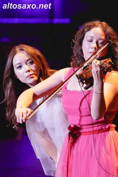 two girls in pink dresses playing violin on stage with purple lighting behind them and the caption altosaxo net