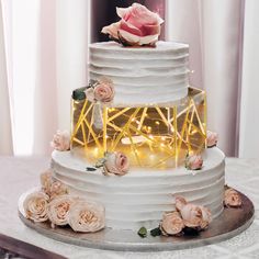 a white wedding cake with pink flowers and lights on the top tier is surrounded by greenery