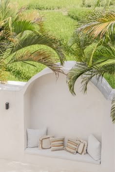 a white bench with pillows on it under a palm tree in front of a grassy field