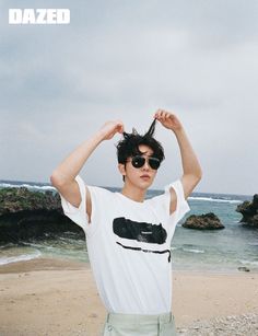 a young man standing on top of a beach next to the ocean holding his hair