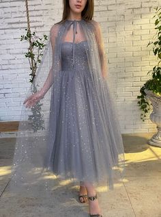 Details:- Starry tulle fabric in a pastel grey hue- Hearty corset top, with built-in pads and boning- Draped bodice- Cinched waist- Inseam pockets- Midi-length skirt- Decorative cape, gathered at the neck and with a tied spaghetti bow-Opti... Dress With Cloak, Prom Dress Glitter, Prom Dresses 2022, Prom Dresses Cheap, Capelet Dress, Vintage Prom Dress, Popular Prom Dresses, Grey Prom Dress