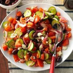 a white plate topped with cucumbers, tomatoes and other veggies next to a bowl of olives