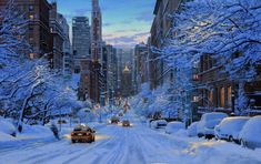 cars driving down a snow covered street in the middle of winter with tall buildings on either side