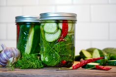 pickles and cucumbers in mason jars on a wooden table with garlic, pepper, and dill