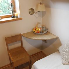 a small room with a bed, table and chair next to the window that has a bowl of fruit on it