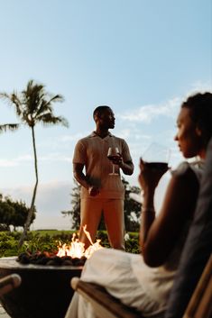 two people sitting around a fire pit with drinks in front of them and palm trees