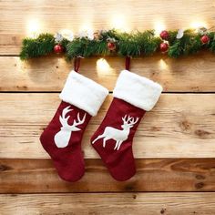 two christmas stockings hanging on a wooden wall with lights around them and an evergreen garland