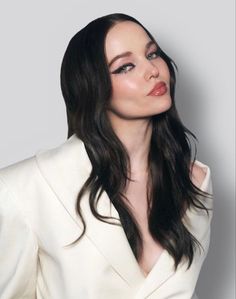 a woman with long black hair wearing a white suit and posing for the camera in front of a gray background