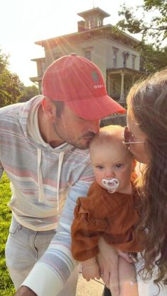 a man and woman holding a baby in front of a house with the sun shining on them