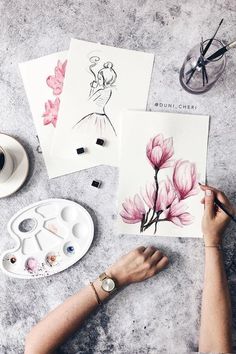 a woman is painting flowers on paper with watercolors and paintbrushes next to it