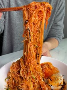 a person holding chopsticks over a bowl of noodles with sauce and vegetables on it