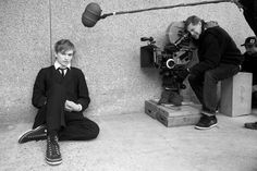 two young men sitting on the ground in front of a camera and another man standing next to them