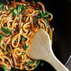 stir fried noodles with meat and vegetables in a wok on the stove top next to a wooden spoon