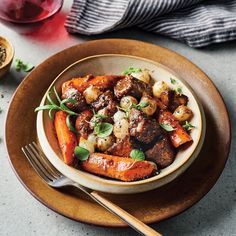 a bowl filled with meat and carrots on top of a wooden plate next to a fork