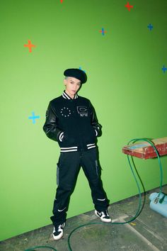 a young man standing in front of a green wall wearing a black jacket and hat