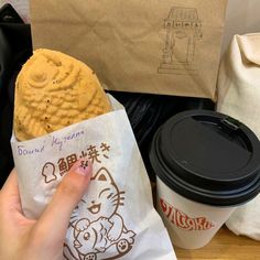 a person holding a cookie in their hand next to a cup of coffee and paper bag