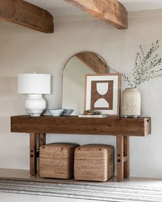 a wooden table topped with baskets under a mirror next to a lamp and vases