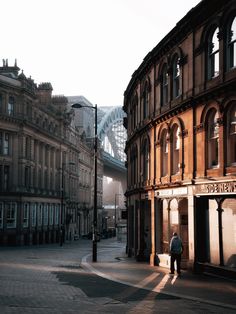 a man is walking down the street in front of an old building with a bridge behind him