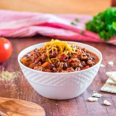 a white bowl filled with chili and cheese on top of a wooden table next to crackers