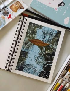 an open notebook on a table with watercolors and other art supplies