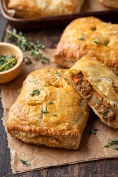 two meat filled pastries on top of a piece of wax paper next to a bowl of herbs