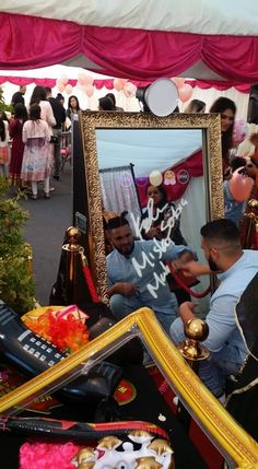 a man sitting in front of a mirror surrounded by other items and people standing around