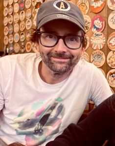 a man wearing glasses and a hat sitting in front of a wall covered with buttons