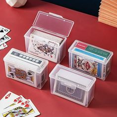 four plastic boxes with playing cards in them on top of a red table next to two stacks of cards