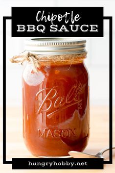 a mason jar filled with barbecue sauce on top of a wooden table next to a spoon