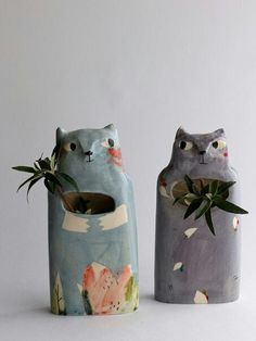 two ceramic vases with plants in them on a white surface, one is shaped like a cat and the other has an animal face