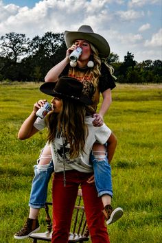 two women in cowboy hats are sitting on a rocking chair and one woman is drinking from a bottle