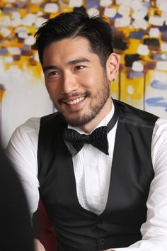 a man wearing a black vest and bow tie sitting in front of a colorful painting