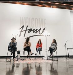 four women sitting on chairs in front of a wall with the words welcome home painted on it