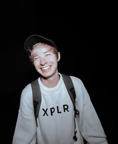 a young man with suspenders and a baseball cap smiles at the camera while wearing a backpack