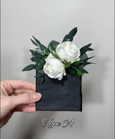 a hand holding a black bag with white flowers in it and green leaves on top