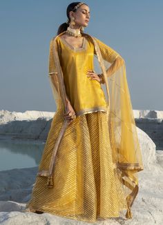 Step into a world of elegance with our Yellow Gota Long Kurta Sharara Set. The shimmer chanderi kurta, adorned with intricate gota work, radiates a regal charm. The matching net sharara mirrors the embellishment, creating a festive look. To complete the look, a light-as-air net dupatta with embellished edges adds an ethereal touch. Perfect for occasions like Haldi, Mehendi, and Puja. Composition : Kurta - Shimmer Chanderi , Sharara and Blouse - Soft Net Care: Dry Clean Only and Vacuum Storage Th Festive Anarkali Gold Sets, Gold Anarkali Style Designer Sharara, Gold Anarkali Style Sharara In Organza, Gold Anarkali Sharara In Organza, Gold Sharara With Resham Embroidery In Chandbali Shape, Gold Sharara With Resham Embroidery For Diwali, Gold Organza Sharara, Semi-stitched Gold Sharara For Designer Wear, Designer Wear Sharara With Gota Work For Festive Season